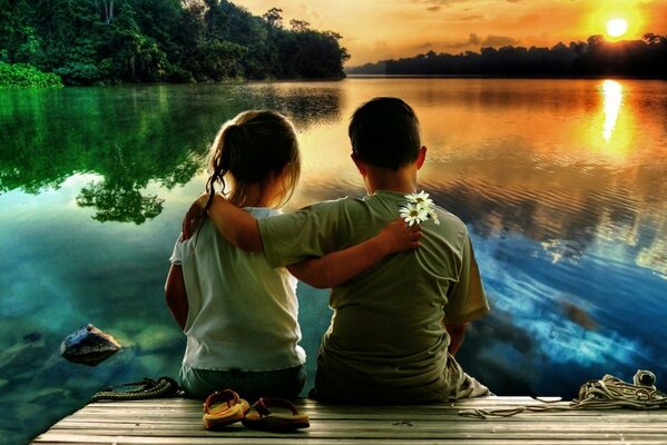 Un niño y una niña se abrazan en la orilla del río y miran la puesta de sol