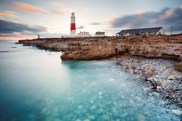 El océano baña la playa rocosa, el faro es visible a lo lejos